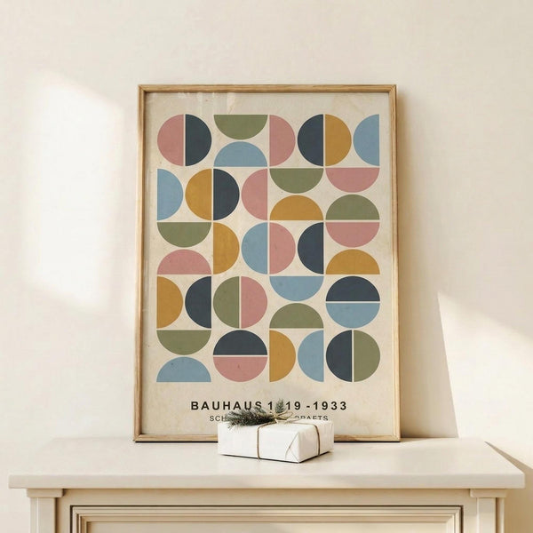 May include: Framed geometric art print with colorful semicircles and quarter-circles in muted tones of pink, blue, green, yellow, and navy arranged in a grid pattern. The poster features "BAUHAUS 1919-1933" text at bottom. Natural wood frame sits on light wooden surface next to small white wrapped gift with greenery sprig against cream wall.
