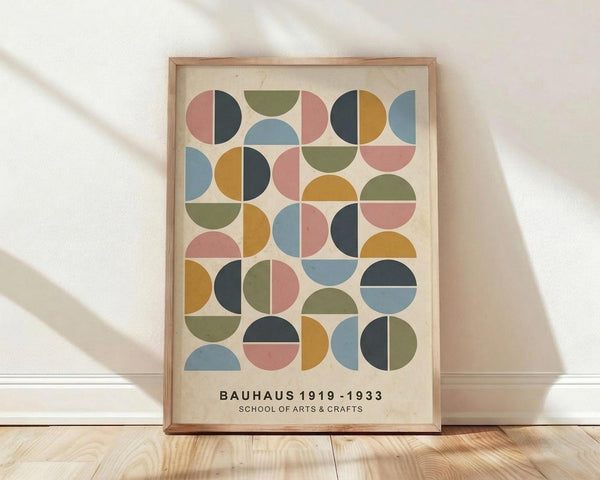 May include: Framed geometric art print featuring colorful circles and semicircles in muted blue, coral pink, olive green, mustard yellow, and navy arranged in a grid pattern on cream background. Text reads "BAUHAUS 1919-1933 SCHOOL OF ARTS & CRAFTS" below the design. Natural wood frame sits on light wooden floor against white wall with soft sunlight shadows.