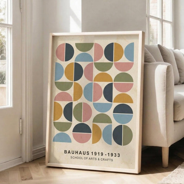May include: A framed poster with geometric pattern of colorful circles and semicircles in muted tones of coral pink, sage green, mustard yellow, dusty blue, and navy arranged in a grid formation on cream background. Light wood frame leaning against white wall beside beige upholstered sofa with throw pillows. Bright natural light streams through tall white-framed windows creating soft shadows on hardwood floors.