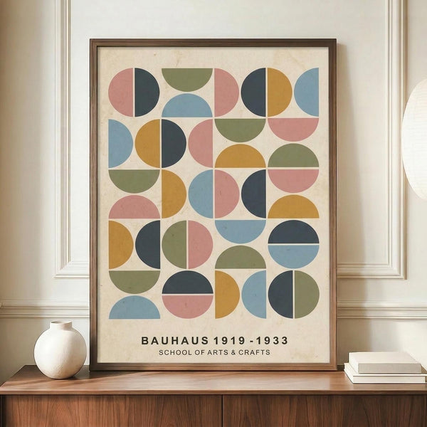 May include: Framed geometric poster with colorful semicircles and circles in muted pink, blue, green, orange and navy arranged in a grid pattern on cream background. Text reads "Bauhaus 1919-1933 School of Arts & Crafts" at bottom. Displayed on wooden sideboard next to small white ceramic vase and stacked books against white paneled wall.