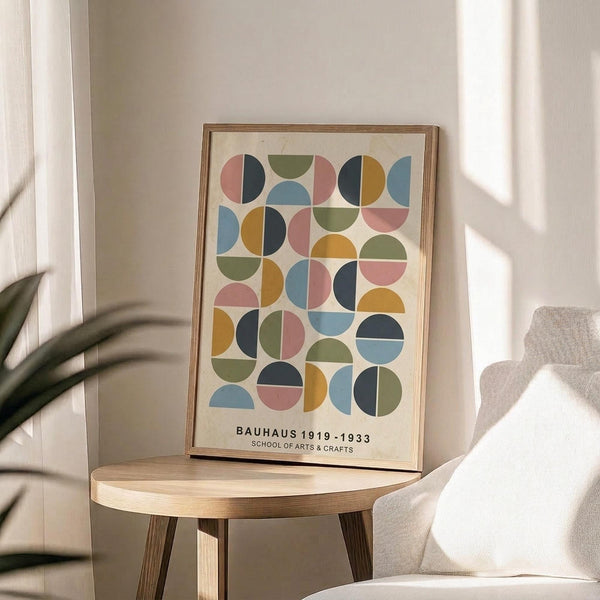 May include: Framed geometric poster with colorful half-circles and crescents in pink, blue, green, orange, and navy arranged in a grid pattern. Text reads "BAUHAUS 1919-1933 SCHOOL OF ARTS & CRAFTS" at bottom. Displayed on round wooden side table next to beige upholstered chair with plant shadows on cream wall in bright natural light.