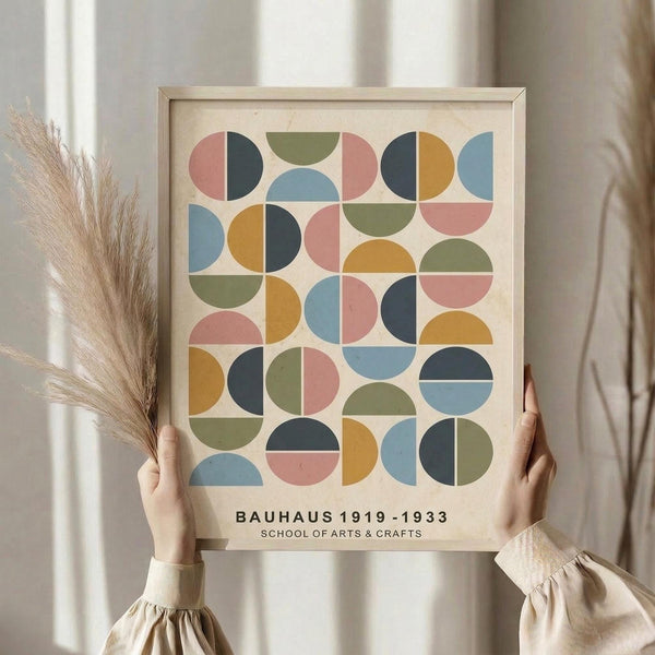 May include: Person holding framed geometric art print featuring colorful semicircles and quarter-circles in muted pink, blue, green, orange and navy arranged in a grid pattern. Light wood frame against cream background with dried pampas grass visible. Bauhaus design with vintage mid-century modern aesthetic displayed in natural lighting near white curtains.