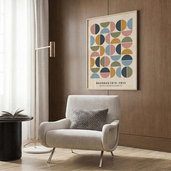 May include: Modern living room mockup showing beige armchair with geometric pillow, gold floor lamp, dark side table with open book, and wood-paneled wall. Framed poster features colorful semicircles in pink, blue, green, yellow, and navy arranged in geometric grid pattern with Bauhaus text below on cream background.