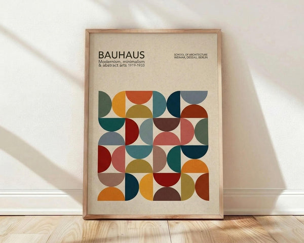 May include: Framed poster with geometric pattern of colorful semicircles and quarter circles in earthy tones including orange, teal, burgundy, olive, and coral on cream background. Text reads "BAUHAUS Modernism, minimalism & abstract arts 1919-1933" and "SCHOOL OF ARCHITECTURE WEIMAR, DESSAU, BERLIN." Natural wood frame leans against white wall on light wooden floor with soft window shadows.