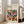May include: Framed geometric art print leaning against white wall next to beige armchair in bright living room. Poster features colorful overlapping semicircles in red, blue, green, orange, and brown arranged in grid pattern on cream background. Black text at top reads "BAUHAUS" with subtitle about modernism and dates. Natural wood frame, approximately 24x30 inches, positioned on light hardwood floor with natural sunlight streaming through tall windows.