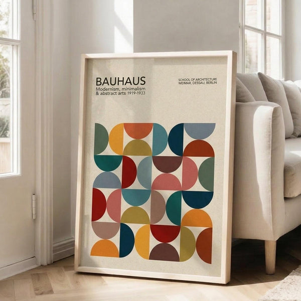 May include: Framed geometric art print leaning against white wall next to beige armchair in bright living room. Poster features colorful overlapping semicircles in red, blue, green, orange, and brown arranged in grid pattern on cream background. Black text at top reads "BAUHAUS" with subtitle about modernism and dates. Natural wood frame, approximately 24x30 inches, positioned on light hardwood floor with natural sunlight streaming through tall windows.