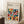 May include: Framed Bauhaus-style poster with colorful geometric semicircles in a grid pattern, featuring vibrant oranges, teals, reds, and earth tones on cream background. Light wood frame leans against white wall next to beige couch, with coffee cup on wooden block nearby on textured rug.
