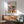 May include: Framed Bauhaus poster with colorful geometric circles in red, orange, blue, green, and brown tones displayed above a wooden mid-century modern credenza. Scene shows mint green upholstered chair with wooden arms, white ceramic table lamp, and round coffee table in a bright room with natural sunlight casting shadows on white walls.