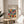 May include: Framed art print with colorful geometric circles and semicircles in a grid pattern, featuring vibrant reds, blues, greens, yellows and earth tones on cream background. The poster sits on a round wooden side table next to a beige upholstered chair, with plant shadows cast on white wall behind. Natural lighting creates soft shadows across the minimalist room setting.