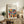 May include: Framed geometric art poster with colorful semicircles and quarter-circles in a grid pattern, featuring red, blue, green, orange, and earth tones. The print sits on a rustic wooden sideboard alongside books, a ceramic vase, and a fern plant. A cream upholstered chair with brown leather cushion is visible in the foreground of this cozy living room setting.