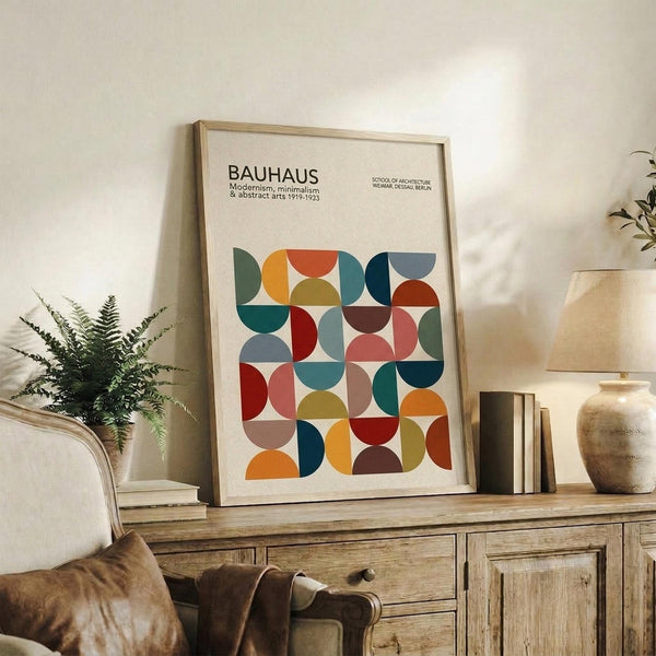 May include: Framed geometric art poster with colorful semicircles and quarter-circles in a grid pattern, featuring red, blue, green, orange, and earth tones. The print sits on a rustic wooden sideboard alongside books, a ceramic vase, and a fern plant. A cream upholstered chair with brown leather cushion is visible in the foreground of this cozy living room setting.