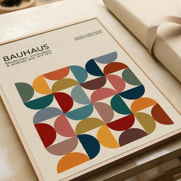 This Bauhaus-inspired poster features overlapping semicircles in vibrant colors including teal, orange, red, yellow, green, and blue arranged in a geometric grid pattern. The minimalist design is printed on cream-colored paper with "BAUHAUS" and "Modernism, minimalism & abstract arts 1919-1933" text at the top. The print is displayed on a wooden surface alongside neutral-toned packaging materials and rolls of paper, creating a warm, artistic workspace setting.