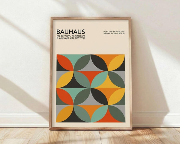 May include: Framed poster with geometric abstract design featuring overlapping circles in orange, yellow, teal, olive green, and black creating a repeating pattern. Text reads "BAUHAUS Modernism, minimalism & abstract arts 1919-1933" and "SCHOOL OF ARCHITECTURE WEIMAR, DESSAU, BERLIN". Light wood frame leaning against white wall on wooden floor with natural shadows.