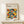 May include: Framed Bauhaus poster with geometric pattern of overlapping circles in orange, yellow, teal, gray, and black colors, displayed on light wood shelf beside small wrapped gift with twine and greenery sprig against cream wall