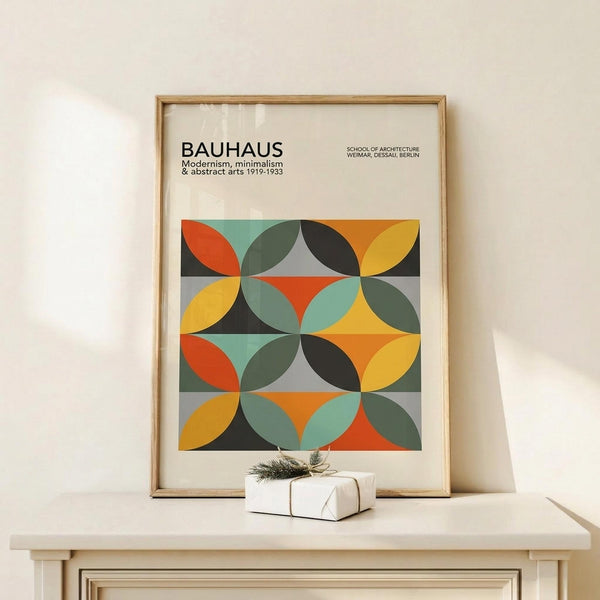 May include: Framed Bauhaus poster with geometric pattern of overlapping circles in orange, yellow, teal, gray, and black colors, displayed on light wood shelf beside small wrapped gift with twine and greenery sprig against cream wall
