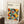 May include: Framed geometric abstract art poster with overlapping circles in orange, yellow, teal, olive green, black and gray creating a modernist pattern. The cream-colored print features "BAUHAUS" text at top. Wooden frame leans against white wall corner next to wooden side table with white coffee cup and beige throw blanket on textured rug.