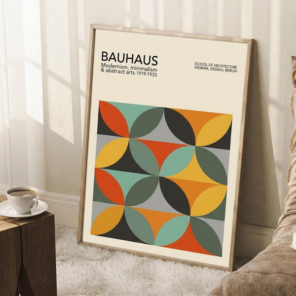 May include: Framed geometric abstract art poster with overlapping circles in orange, yellow, teal, olive green, black and gray creating a modernist pattern. The cream-colored print features "BAUHAUS" text at top. Wooden frame leans against white wall corner next to wooden side table with white coffee cup and beige throw blanket on textured rug.
