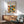 May include: Framed Bauhaus poster with geometric pattern of overlapping circles in orange, green, yellow and black colors, displayed above wooden sideboard with white lamp in modern living room setting with mint green armchair and natural lighting casting shadows on white wall.