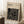 May include: Framed beige poster with black geometric maze-like pattern and "BAUHAUS" text at top, leaning against white wall next to wooden side table with white coffee cup, soft lighting creating shadows across cream-colored textured rug and cozy neutral interior setting.