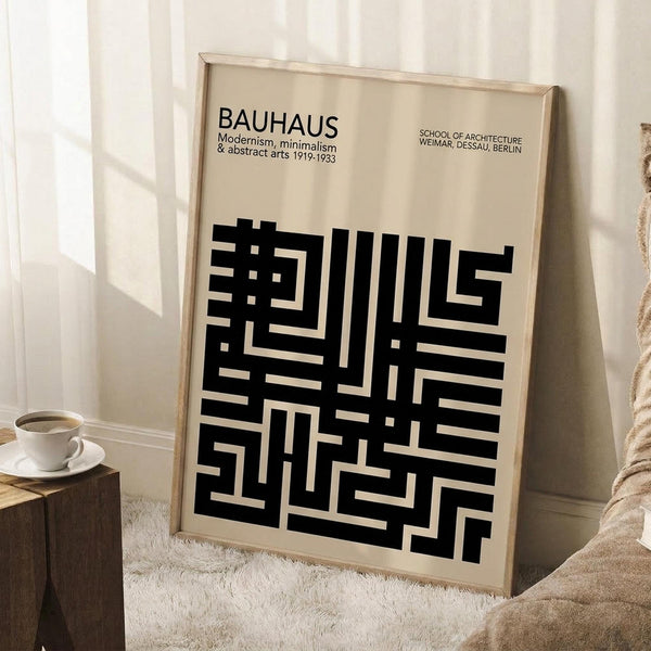 May include: Framed beige poster with black geometric maze-like pattern and "BAUHAUS" text at top, leaning against white wall next to wooden side table with white coffee cup, soft lighting creating shadows across cream-colored textured rug and cozy neutral interior setting.