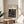 May include: Black and white Bauhaus poster with geometric maze-like pattern displayed in thin wooden frame on round wooden side table next to beige upholstered chair. Soft natural lighting creates shadows on cream wall behind, with green plant leaves visible in corner of minimalist modern room setting.