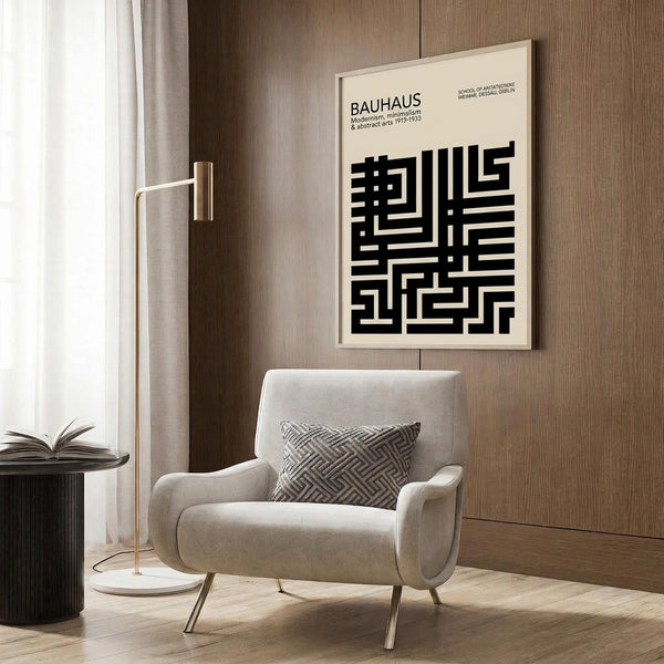 May include: Beige armchair with geometric patterned pillow positioned near wood paneled wall. Black and white geometric poster in light frame displays Bauhaus text and abstract maze-like design. Gold floor lamp with white base stands beside chair. Dark round side table with open book visible on left. Neutral curtains frame window providing natural light.