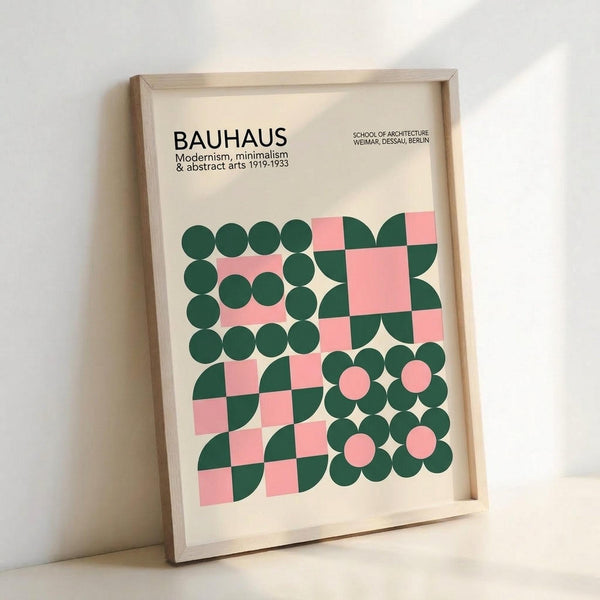 May include: Framed Bauhaus-inspired poster with geometric design featuring dark green circles and pink squares arranged in an abstract pattern. The cream-colored print displays "BAUHAUS" text at top with "Modernism, minimalism & abstract arts 1919-1933" and "School of Architecture Weimar, Dessau, Berlin" below. Natural wood frame leans against white wall on light surface, showing clean minimalist styling typical of Bauhaus movement.