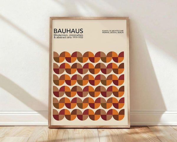 May include: Framed Bauhaus poster with geometric pattern of overlapping circles in warm autumn colors - oranges, browns, burgundy, and rust tones arranged in rows. The minimalist design features text about the German art school from 1919-1933. Natural wood frame sits against white wall on light wooden floor with soft shadows from window light.