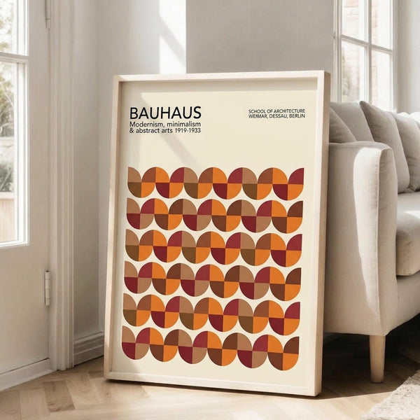 May include: Framed Bauhaus poster with geometric pattern of overlapping circles in warm autumn colors - oranges, browns, and burgundy - leaning against white wall next to beige upholstered chair in bright, minimally decorated room with large windows and natural light streaming across wooden floors.