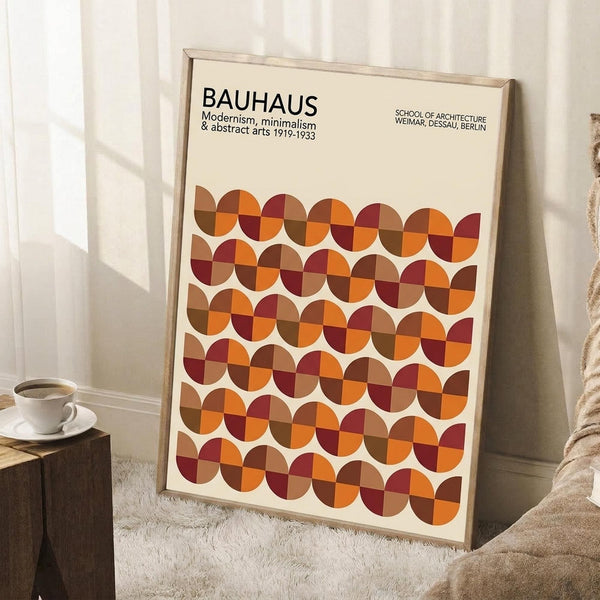 May include: Framed Bauhaus-style poster with geometric pattern of overlapping circles in warm autumn colors - orange, burgundy, brown, and tan - leaning against white wall. Cream-colored frame, positioned next to wooden side table with white coffee cup and saucer, cozy beige throw blanket visible. Clean minimalist room setting with natural lighting creating soft shadows on textured area rug.