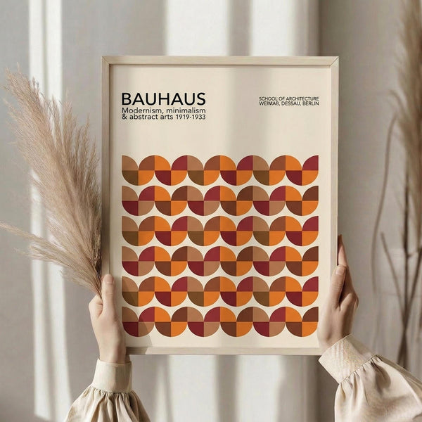 May include: Person holding framed Bauhaus poster featuring geometric pattern of overlapping circles in warm autumn colors - orange, burgundy, brown, and rust red tones. Light wood frame against cream background with dried pampas grass visible to the left. Clean minimalist styling with person wearing beige knit sleeves.