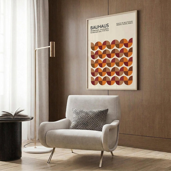 May include: Framed Bauhaus poster with geometric semicircle pattern in warm orange, burgundy, and brown tones hanging on wood-paneled wall above beige upholstered armchair with geometric throw pillow. Modern floor lamp with brass finish and marble base stands nearby next to round black side table with open book. Neutral interior with curtains and light wood flooring creates minimalist mid-century modern aesthetic.