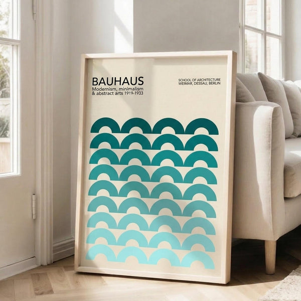 May include: Framed Bauhaus poster with geometric turquoise and teal semicircle pattern leaning against white wall next to beige armchair. Natural sunlight streams through windows onto light wooden floors. The minimalist design features overlapping arched shapes in gradient blues on cream background within thin wooden frame.