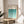 May include: Framed Bauhaus poster with geometric teal and turquoise semicircle pattern in gradient ombre effect, displayed on light wood round side table next to beige upholstered chair. Natural sunlight casts window shadows on cream wall behind the minimalist modernist artwork in thin wooden frame.