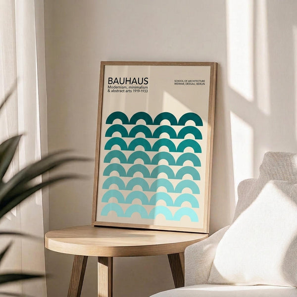 May include: Framed Bauhaus poster with geometric teal and turquoise semicircle pattern in gradient ombre effect, displayed on light wood round side table next to beige upholstered chair. Natural sunlight casts window shadows on cream wall behind the minimalist modernist artwork in thin wooden frame.