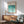 May include: Framed Bauhaus poster with teal gradient wave pattern displayed above mid-century modern wooden credenza. Sage green upholstered armchair with wooden frame sits nearby, along with white ceramic table lamp. Warm natural lighting creates diagonal shadows on white wall, showcasing minimalist interior design with vintage furniture pieces.