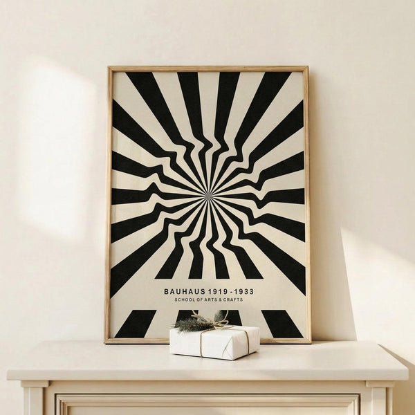 May include: Black and white optical illusion poster with radiating wavy stripes creating a hypnotic sunburst pattern, displayed in light wood frame on cream-colored wooden surface next to small wrapped white gift box with twine and greenery sprig against neutral wall