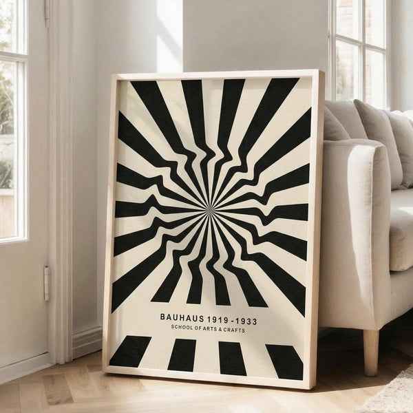 May include: Black and white geometric poster with radiating sunburst pattern creating an optical illusion effect, displayed in light wood frame leaning against white wall next to beige upholstered sofa. The design features bold alternating stripes emanating from center point with wavy distortion. Text at bottom reads "BAUHAUS 1919-1933 SCHOOL OF ARTS & CRAFTS" in simple black lettering on cream background.