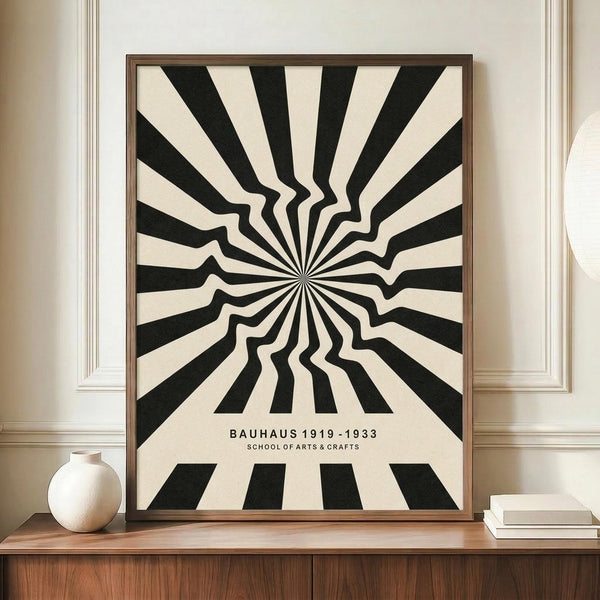 May include: Black and white optical illusion poster with radiating zigzag pattern creating tunnel-like depth effect, displayed in thin brown frame on wooden sideboard. Text reads "BAUHAUS 1919-1933 SCHOOL OF ARTS & CRAFTS" below the geometric design. Small white ceramic vase and stacked books sit nearby on warm wood surface against cream walls with white trim molding.
