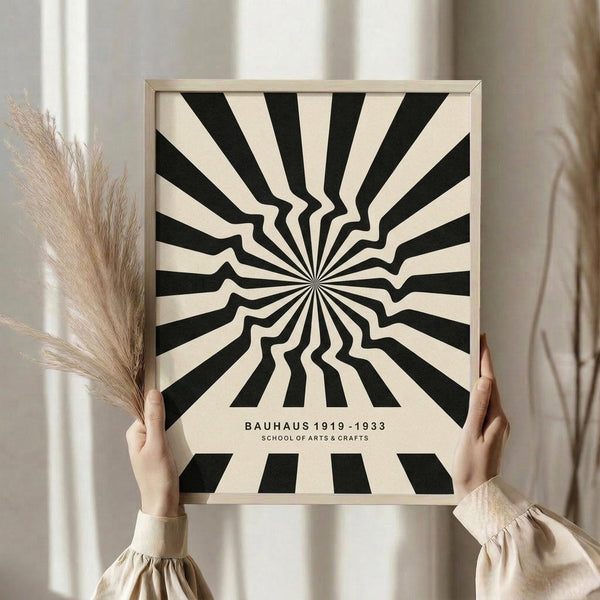 May include: Person holding framed black and cream optical illusion print featuring radiating wavy stripes emanating from center point, with "BAUHAUS 1919-1933 SCHOOL OF ARTS & CRAFTS" text below. Light wood frame, neutral beige clothing, dried pampas grass visible, soft natural lighting against white curtained background.