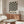 May include: Black and white optical illusion poster with radiating wavy stripes creating a hypnotic sunburst pattern, displayed above a mid-century modern walnut credenza in a minimalist living room. The framed artwork features text at bottom, flanked by a white sculptural chair and brass floor lamp, with a glass-top oval coffee table in foreground.