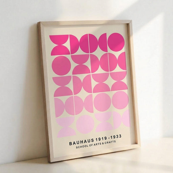 May include: Framed geometric art print leaning against white wall, featuring pink circles and semicircles in gradient shades from bright magenta to pale pink arranged in a grid pattern, with "BAUHAUS 1919-1933 SCHOOL OF ARTS & CRAFTS" text at bottom, displayed in light wooden frame on cream background