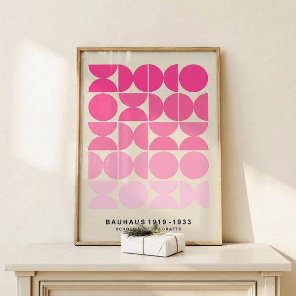 May include: Framed geometric art print with pink circular and semi-circular shapes arranged in a grid pattern, displayed on a light wood console table. The poster features a gradient from bright fuchsia at top to pale pink at bottom with "BAUHAUS 1919-1933 SCHOOL ARTS CRAFTS" text. A small white wrapped gift sits beside the natural wood frame against a cream wall.