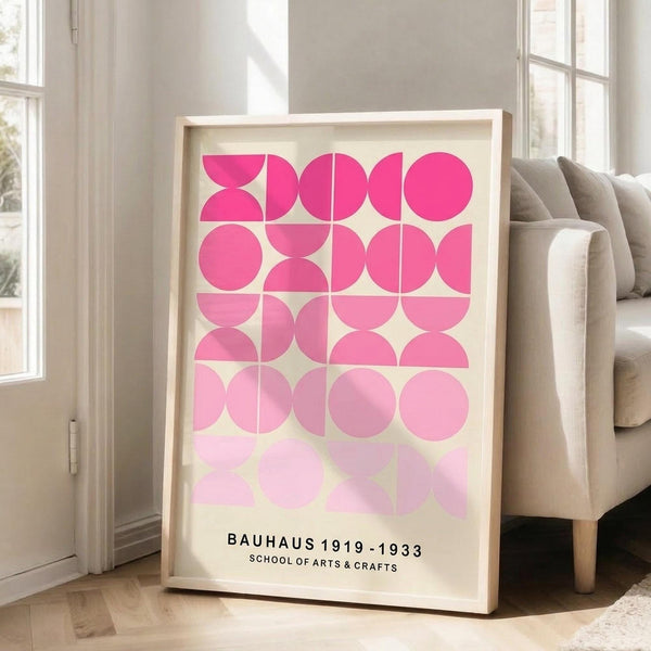 May include: A framed geometric art print with pink circular and semi-circular shapes arranged in a grid pattern on cream background, displayed in light wood frame leaning against white wall next to beige upholstered sofa in bright, naturally-lit room with large windows and hardwood floors.