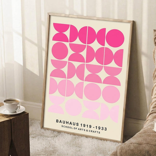 May include: Framed geometric poster with pink and white circular shapes in a grid pattern, leaning against white wall corner. Light wood frame, cream background with black text at bottom. Coffee cup on wooden block nearby, beige textile visible, soft natural lighting creating shadows.