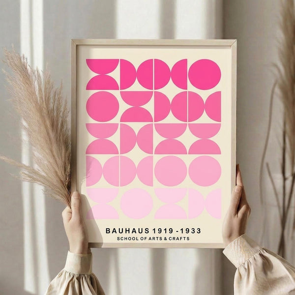 May include: Hands holding a framed geometric art print with pink gradient circles and semicircles in a grid pattern, text reading "BAUHAUS 1919-1933 SCHOOL OF ARTS & CRAFTS" at bottom. Light wood frame against cream background with dried pampas grass visible on left side. Person wearing beige ruffled sleeve top.