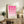 May include: Framed pink geometric poster with gradient circles and semicircles in a wooden frame, displayed on rustic wood sideboard alongside fern plant, ceramic vases, and stacked books in neutral-toned living room setting with beige chair visible.