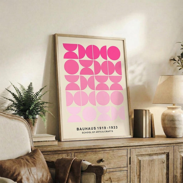 May include: Framed pink geometric poster with gradient circles and semicircles in a wooden frame, displayed on rustic wood sideboard alongside fern plant, ceramic vases, and stacked books in neutral-toned living room setting with beige chair visible.