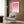 May include: Framed geometric poster with pink gradient circles and semicircles arranged in a grid pattern, displayed on warm wood paneled wall above a cream upholstered armchair with geometric throw pillow. Modern brass floor lamp and dark side table with open book complete the contemporary living room setting with natural light from window.