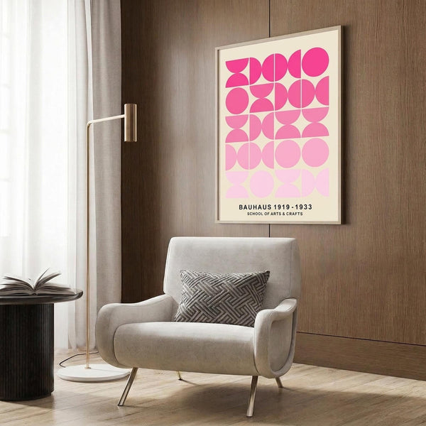 May include: Framed geometric poster with pink gradient circles and semicircles arranged in a grid pattern, displayed on warm wood paneled wall above a cream upholstered armchair with geometric throw pillow. Modern brass floor lamp and dark side table with open book complete the contemporary living room setting with natural light from window.